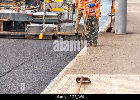 Ein Industriefertiger legt eine Schicht neuen heißen Asphalts auf die Fahrbahn. Der Arbeiter kontrolliert die Ebenheit des Pflasterbodens mit einer Schaufel. Speicherplatz kopieren. Stockfoto