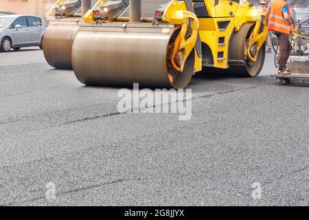 Die Metallzylinder der gelben Schwingwalze verdichten den frischen Asphalt kraftvoll auf dem neuen Straßenbau. Platz zum Kopieren, Nahaufnahme. Stockfoto