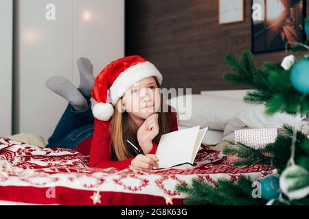 Das rothaarige, niedliche Teenager-Mädchen in einem Weihnachtshut schreibt einen Brief an den Weihnachtsmann. Stockfoto