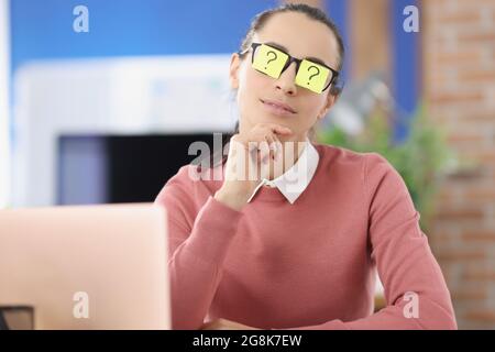 Im Büro sitzt eine junge Frau mit Aufklebern mit Fragezeichen auf der Brille Stockfoto
