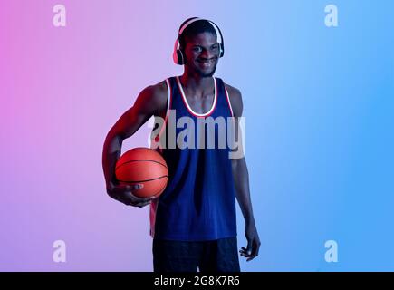 Fröhlicher schwarzer Basketballspieler, der Musik in Kopfhörern genießt, den Ball hält und in Neonbeleuchtung lächelt Stockfoto