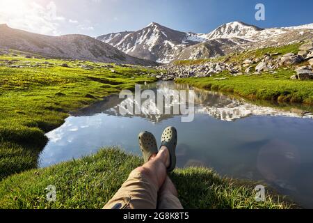 Die Beine des Wanderers in grünen Schuhen ruhen in der Nähe des klaren Bergsees mit Spiegelung des Gipfels mit Schneekappe Stockfoto