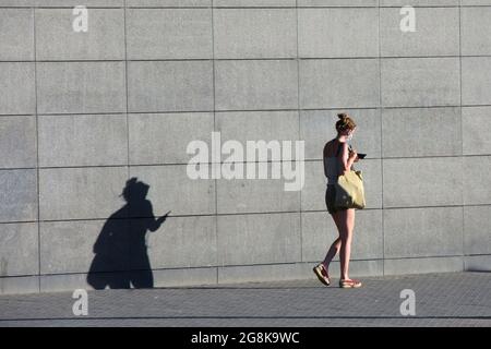 Die junge Frau geht an einer grauen Wand entlang. Sie trägt Sommerkleidung. Sie läuft mit Maske, während sie ihr Smartphone mit Kopfhörern hört. Stockfoto