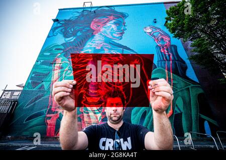 Der Organisator von 'Upfest' Steve Hayles hält ein rotes Filtergel neben einem Graffiti-Stück des Künstlers Insane 51, das ein interaktives Stück ist und sich durch einen blauen oder roten Filter, wie die in 3D-Brillen, anders betrachten lässt. Eine Frau oder das Skelett in ihrem Körper wird an einer riesigen Wand in Bedminster, Bristol, enthüllt, während das Wandgemälde-Projekt von 75 den halben Punkt erreicht und im Sommer verschiedene Graffiti-Stücke auf Wände und Gebäude in der ganzen Stadt gesprüht werden. Bilddatum: Mittwoch, 21. Juli 2021. Stockfoto