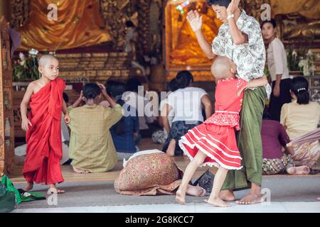 Der junge burmesische Novizenmönch sieht so aus, als würde ein junges Mädchen mit rasiertem Kopf ihren Vater, die Shwedagon Pagode, Yangon, Myanmar, umarmen Stockfoto