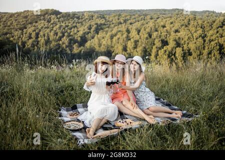 Menschen- und Freundschaftskonzept. Fröhliche Gesellschaft von drei Freundinnen, Spaß haben und genießen Sommer-Picknick und Toasting Gläser mit Wein, machen Selfie-Foto auf dem Smartphone Stockfoto