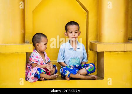 Der junge Burmesin sitzt in Lotusposition und meditiert, während die jüngere Schwester Shwedagon Pagode, Yangon, Myanmar, anschaut Stockfoto