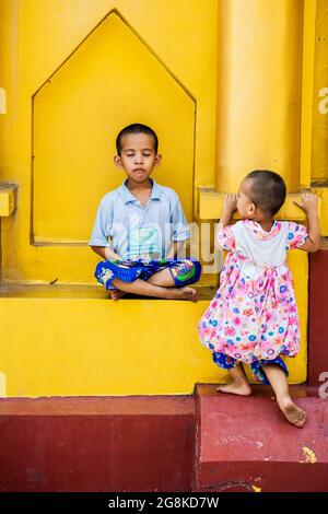 Der junge Burmesin sitzt in Lotusposition und meditiert, während die jüngere Schwester Shwedagon Pagode, Yangon, Myanmar, anschaut Stockfoto