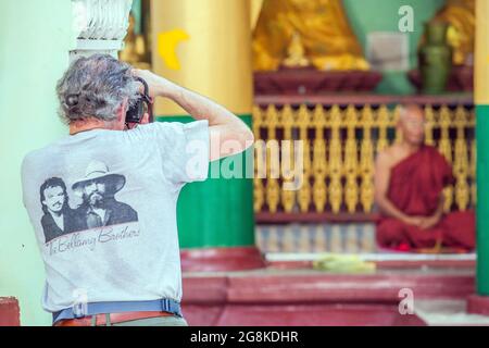 Westlicher Tourist mit T-Shirt von Bellamy Brothers fotografiert einen älteren Mönch (unscharf), der in Lotusposition im Shwedagon Tempel, Yangon, Myanmar saß Stockfoto