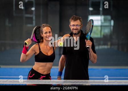 Portrait von zwei lächelnden Sportlern, die in der Halle auf dem Padel Court mit Schlägern und Bällen posieren Stockfoto
