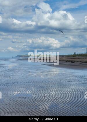 Ein Blick auf Redcar Beach, mit dem Meer und benetztem Sand, der die verschlungenen Wolken eines sommerlichen Himmels und das sonnenbeschienene Hunt Cliff am Horizont reflektiert. Stockfoto