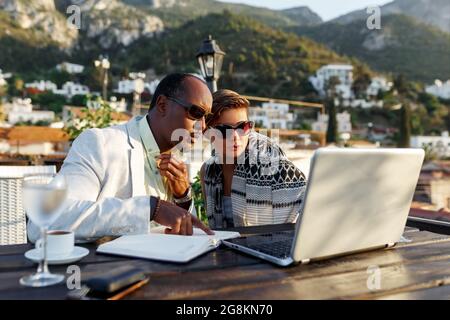 Coworking und freiberufliches Konzept. Gemischtes Rennen, schwarze Haut im mittleren Alter Mann und Frau arbeiten zusammen am Laptop, während sie auf der Cafeterrasse sitzen. Stockfoto