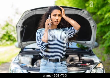 Nervöse Frau mit offener Autohaube, die den Autoservice anruft Stockfoto