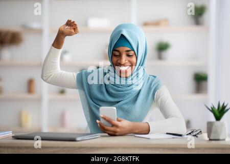 Überglücklich schwarze muslimische Frau in Hijab Blick auf Smartphone und feiert Erfolg Stockfoto