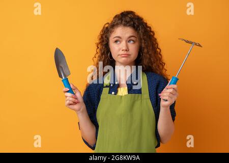 Junge Gärtnerin Frau trägt Schürze hält Gartengeräte vor gelbem Hintergrund Stockfoto