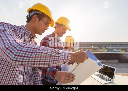 Diverse Gruppe von drei Ingenieuren, Caucacian und Asian, auf der Baustelle, die sich den Bauplan und das Brainstorming während der Arbeit auf dem Dach des Gebäudes angeschaut haben Stockfoto