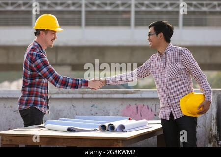 Zwei Ingenieure, Asiaten und Kaukasierinnen, die nach der Arbeit auf dem Dach der Baustelle mit schönen Sonnenlichtern im Hintergrund die Hände schütteln. Stockfoto