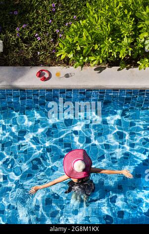 Die Frau trägt einen großen Hut, die Arme ausgebreitet und geht im Pool zu einem Glas Orangensaft und roten Kopfhörern. Stockfoto
