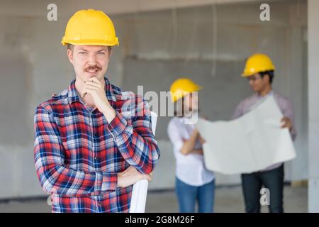 Kaukasischer Ingenieur mit gelbem Sicherheitshut im Stehen und Blick zur Kamera. Es gibt zwei Ingenieure, die im Hintergrund unscharf sprechen. Stockfoto
