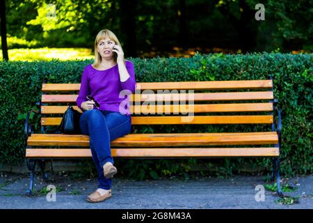 Unschärfe kaukasisch blonde Frau reden, sprechen am Telefon draußen, im Freien. 40-jährige Frau in lila Bluse im Park auf der Bank. Erwachsene Frauen usin Stockfoto