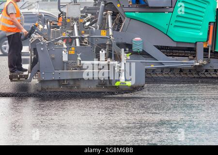 Ein Straßenarbeiter betreibt einen Asphaltfertiger und gießt Asphaltbeton, während er an einem Sommertag hinter einem Bedienfeld auf einer Metallstufe steht. Stockfoto