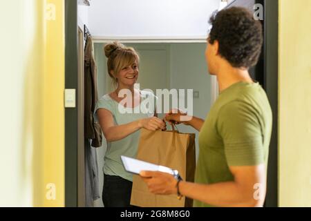 Eine fröhliche und lächelnde Frau an der Tür ihrer Wohnung holt die Lieferung in einer Papiertüte von einem Mann mit Lebensmittelzustellung ab. Stockfoto