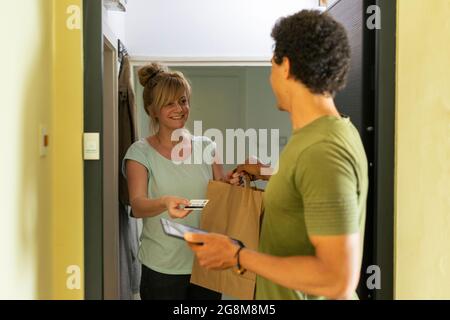 Eine glückliche und lächelnde Frau an der Tür ihrer Wohnung holt die Lieferung in einer Papiertüte von einer Zustellerin ab, die sie mit einer Kreditkarte bezahlt. Stockfoto