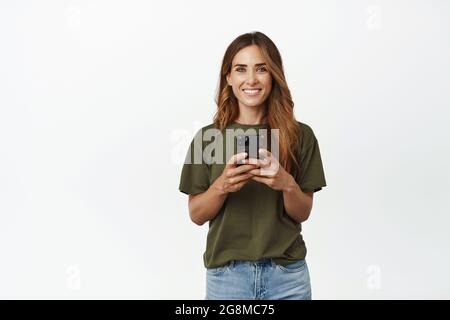 Lächelnde Brünette Frau mit Handy-App, hält Smartphone und schaut glücklich auf die Kamera, stehend in T-Shirt und Jeans vor weißem Hintergrund Stockfoto