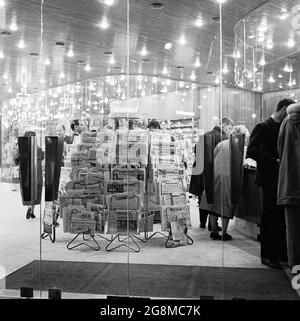 In einem gut beleuchteten, glitzernden Einzelhandelsgeschäft mit einem großen Glaseingang auf der Avenue des Champs-Elysees, in dem sich vor Paris, Frankreich, eine Ausstellung oder ein Regal mit französischen Tageszeitungen befinden, stöbern die Käufer aus den 1950er Jahren, der Vergangenheit, der Abendzeit und aus Paris. Stockfoto
