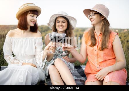 Junggesellinnenabschied. Mädchen Freundschaft und Spaß. Gruppe von drei jungen Frauen in Hüten und Kleidern, genießen leckeres Essen, Wein bei Sommerfest Picknick, klirrende Gläser mit Rotwein. Stockfoto