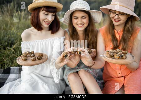 Ästhetisches Picknick im Freien mit Gourmet-Food-Schnecken. Drei hübsche lächelnde weibliche beste Freundinnen, die auf der Wiese auf der Decke sitzen und Holzteller mit leckeren Escargots und Schnecken in der Hand halten Stockfoto