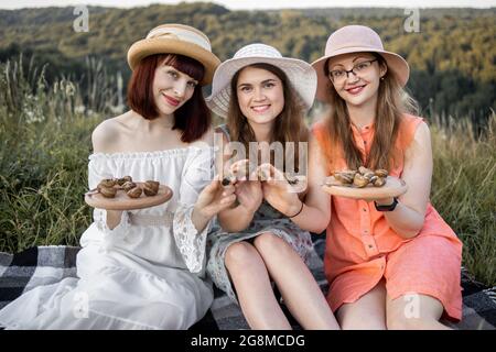 Ästhetisches Picknick im Freien mit Gourmet-Food-Schnecken. Drei hübsche lächelnde weibliche beste Freundinnen, die auf der Wiese auf der Decke sitzen und Holzteller mit leckeren Escargots und Schnecken in der Hand halten Stockfoto