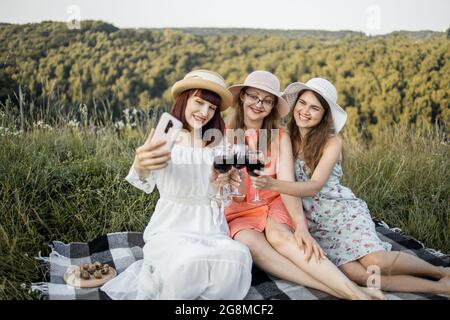 Menschen- und Freundschaftskonzept. Fröhliche Gesellschaft von drei Freundinnen, Spaß haben und genießen Sommer-Picknick und Toasting Gläser mit Wein, machen Selfie-Foto auf dem Smartphone Stockfoto