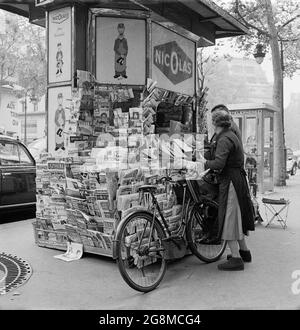 1950er Jahre, historisches Bild von zwei Personen, eine Dame mit ihrem Fahrrad draußen an einem Zeitungskiosks im Zentrum von Paris, Frankreich. Sie kamen erstmals 1857 zum Einsatz, als Baron Haussmann die Pariser Landschaft renovieren wollte. Der Kiosk ist voll von Zeitungen, der Grund dafür ist, dass nach französischem Recht alle Zeitungstitel getragen werden mussten, sodass alle politischen Meinungen zu sehen waren. Auf dem Kiosk wird für Nicolas, das seit langem etablierte pariser Weinunternehmen, Werbung gemacht. Stockfoto