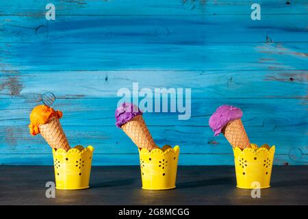 Drei Eiszapfen auf altem, blauem Holzhintergrund. Die Kugeln sind aus Heidelbeere, Erdbeere und Aprikoseneis in Waffeln sortiert Stockfoto