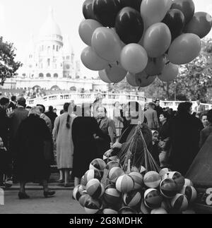 1950, historisches Bild einer Ballonverkäuferin mit ihren Waren, die unter den Besuchern der Basilika des Heiligen Herzens von Paris, besser bekannt als Sacre-Coeur, einer römisch-katholischen Kirche, die auf dem Hügel von Montmartre, Paris, Frankreich, erbaut wurde, herumläuft. Dieses ikonische Denkmal wurde 1919 geweiht und ist das am zweithäufigsten besuchte religiöse Gebäude in Paris. Neben luftgefüllten Ballons hat der Verkäufer auch leichte Strandbälle zum Verkauf. Stockfoto