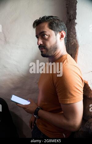 Gutaussehender bärtiger hispanischer Mann mit einem orangefarbenen T-Shirt und einem Smartphone Stockfoto