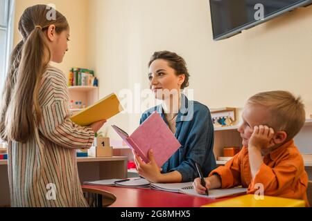 Mädchen mit Buch, das in der Nähe des Lehrers steht, und Junge, der im Klassenzimmer am Schreibtisch sitzt Stockfoto