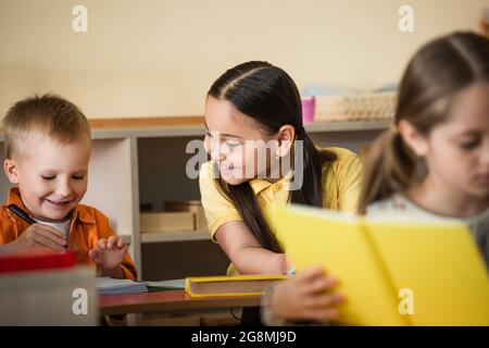 Fröhliches asiatisches Mädchen, das Freund während des Unterrichts in der montessori-Schule in der Nähe des Mädchens auf verschwommenem Vordergrund ansieht Stockfoto