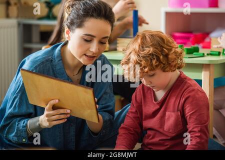 Junge Lehrerin, die während des Unterrichts einem Rotschopf-Jungen ein Holzbrett zeigt Stockfoto