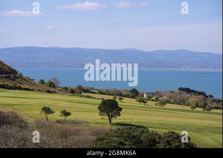 Landschaftlich schöner, heller Blick auf den Golfplatz mit Bäumen, das irische Meer, die Bucht von Dublin, den Leuchtturm von Poolbeg und die Dublin Mountains von Howth, Dublin aus gesehen Stockfoto