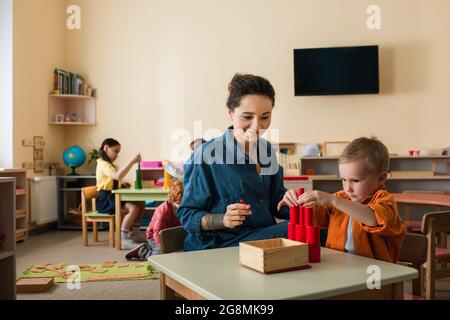 Junge Lehrerin lächelt in der Nähe des Jungen, der aus Holzzylindern in der Nähe von Kindern einen Turm auf unscharfem Hintergrund macht Stockfoto