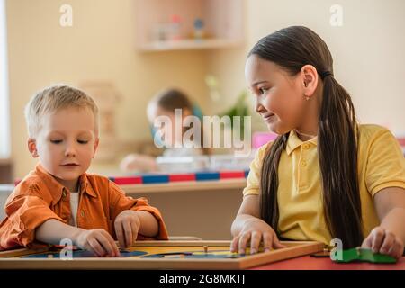 asiatisches Mädchen lächelt, während die Kombination Weltkarte Puzzle mit Jungen in montessori Schule Stockfoto