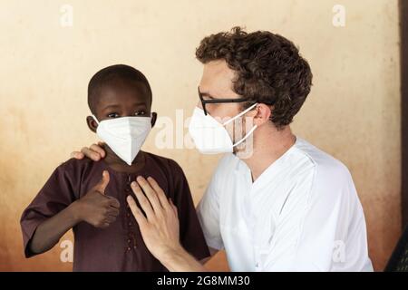 Weißer Doktor hilft afrikanischem schwarzen Jungen. Beide tragen Gesichtsmasken, um die Ausbreitung der anhaltenden globalen Pandemie zu verhindern. Stockfoto