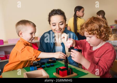 Junge Lehrerin lächelt neben Jungen, die mit bunten Würfeln spielen, und interracial Mädchen auf verschwommenem Hintergrund Stockfoto