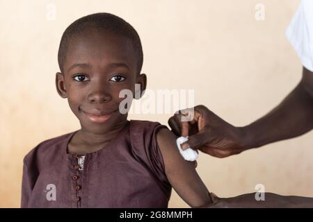 Auf diesem Bild reinigt ein schwarzer Arzt die Injektionsstelle mit einem Wattebausch, nachdem er einen niedlichen lächelnden afrikanischen Prescan geimpft hat Stockfoto