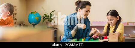 montessori-Lehrerin zeigt auf bunte Blöcke, während sie mit einem asiatischen Mädchen, einem Banner, spielt Stockfoto