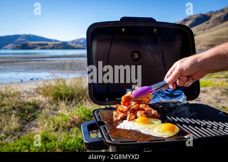 Ein Mann kocht Speck und Eier auf einem tragbaren grill am Meer an einem sonnigen Wintertag, der durch Neuseeland reist Stockfoto