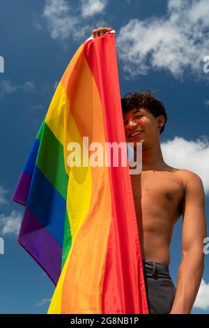 Porträt eines jungen schwulen hispanischen Jungen, der die Kamera anschaut und die LGBT-Flagge hält – Fokus auf vertikales Bild im Gesicht Stockfoto