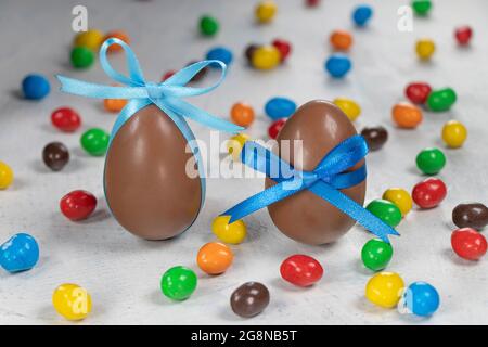 Zwei große Schokoladeneier mit blauen Schleifen und Ohrbändern auf einem Hintergrund aus bunten Süßigkeiten. Weicher Fokus Stockfoto